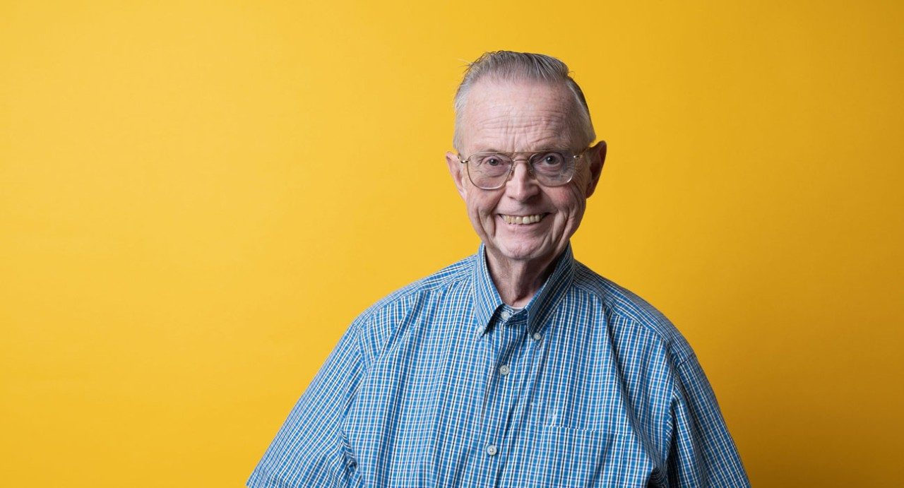 Caregiver and donor William Brady stands in front of a yellow background, smiling. He's a man in his 70s, wearing glasses and a blue button-down shirt.