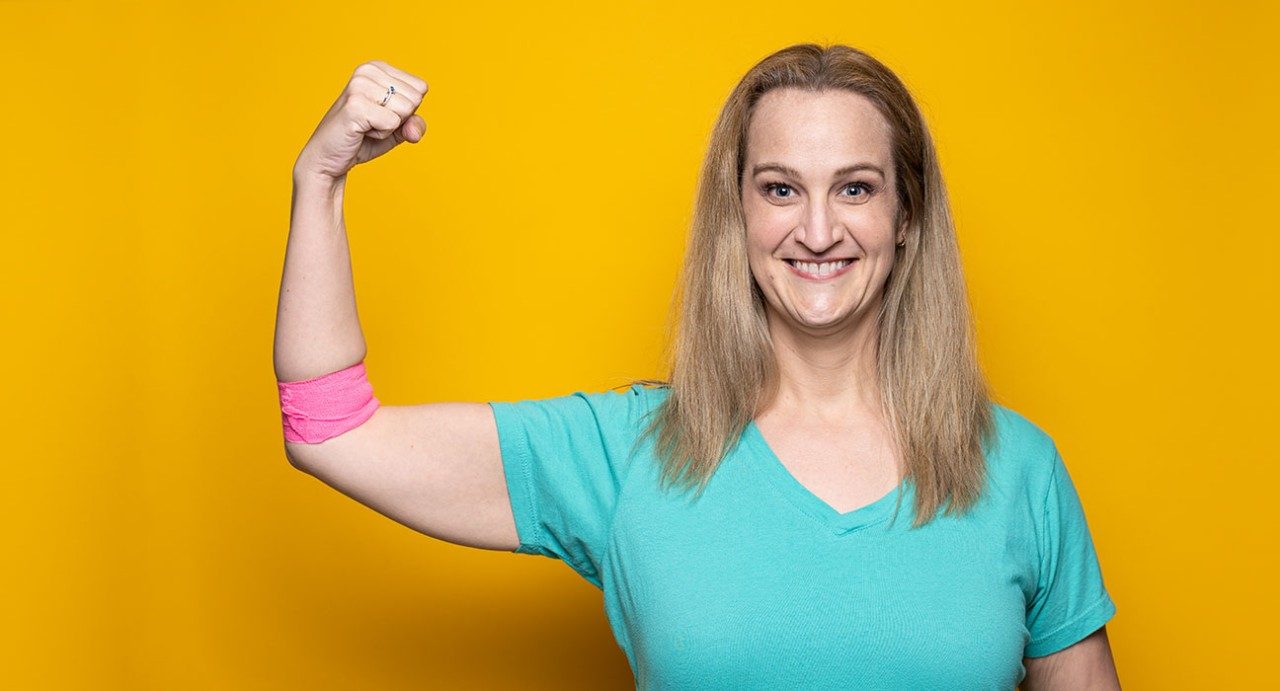 Bile duct cancer caregiver and blood donor Melissa Fuquay Baker stands in front of a yellow background while holding up her arm to show the pink bandage after donating blood.