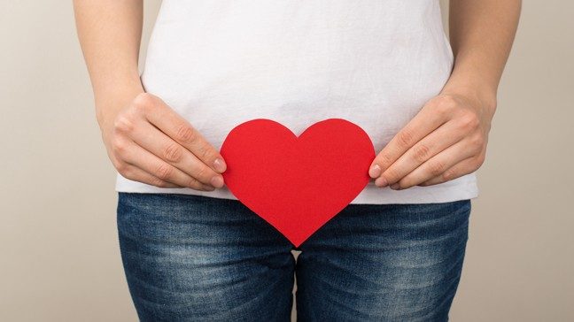Close up of woman's hands holding a bright red paper heart in front of her pelvis