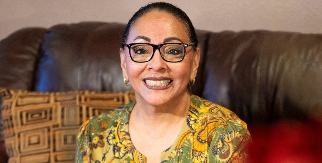 A smiling woman in a yellow and green blouse sits on a brown couch.