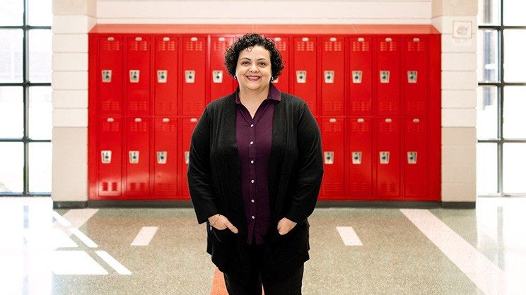 A brunette woman wearing a purple shirt and black cardigan stands in a hallway with a row of red metal lockers behind her