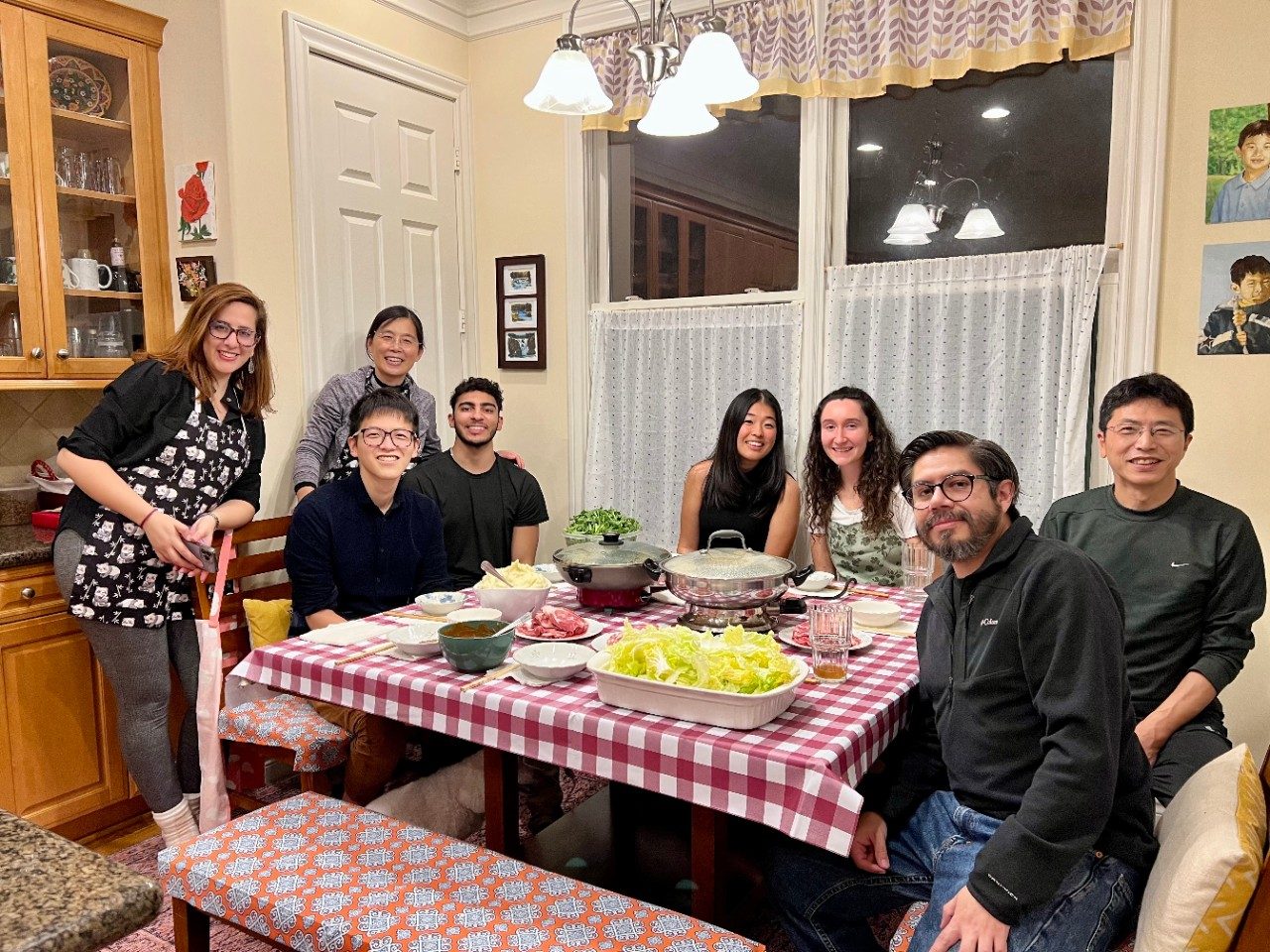 Eight people gathered around a square table with a red and white checkered tablecloth and hot pot ingredients on the table