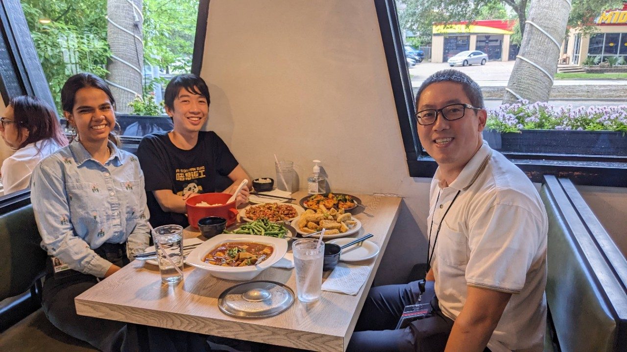 Three people seated around a rectangular table full of food.